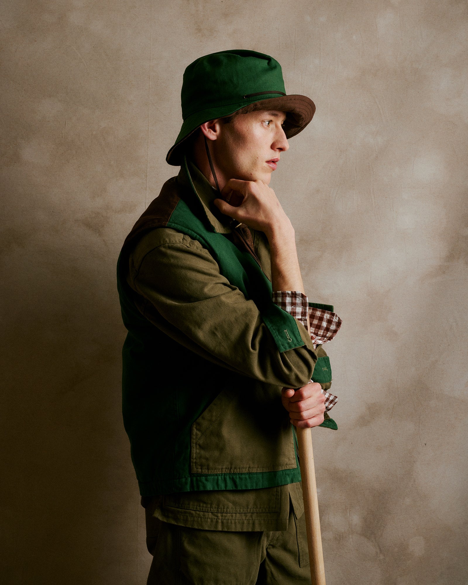 Rovi Lucca FW24 Orto Botanico – Model wearing a quilted gillet in 100% cotton canvas with tones of Bottle, Khaki, and Brown, a khaki unlined garden jacket in 100% cotton canvas with contrast details in Bottle and Brown, khaki 100% cotton garden pants, a brown and panna vichy 100% cotton flannel shirt with contrasting Bordeaux pocket, a 100% Shetland wool Loden neck tie, and a bucket hat.